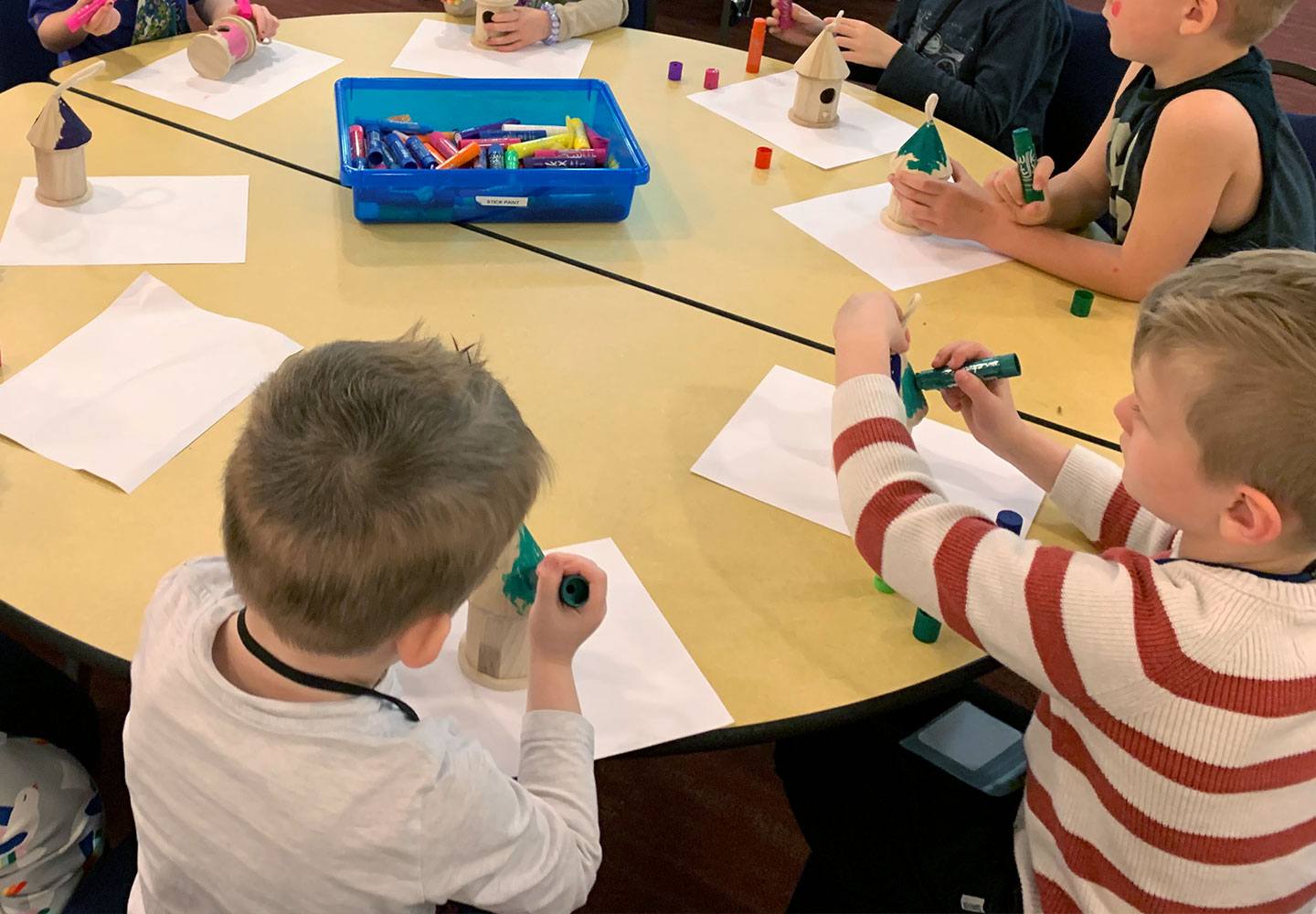 Children are seated around a table, drawing on paper and decorating small wooden shapes with markers. A blue tray filled with markers is in the center of the table.