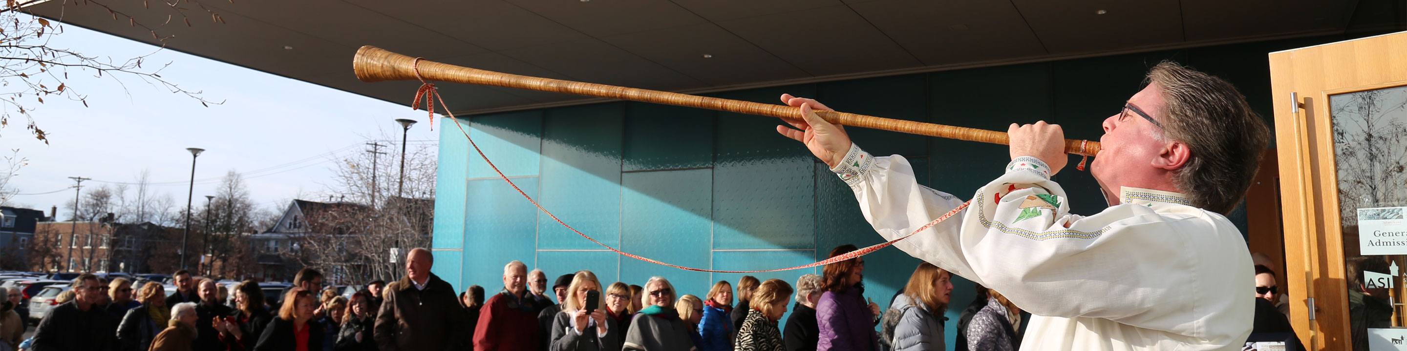 A person in traditional attire plays a long wooden alphorn outside a building, drawing attention from a crowd of people gathered nearby. The scene is set in an urban area with trees in the background.