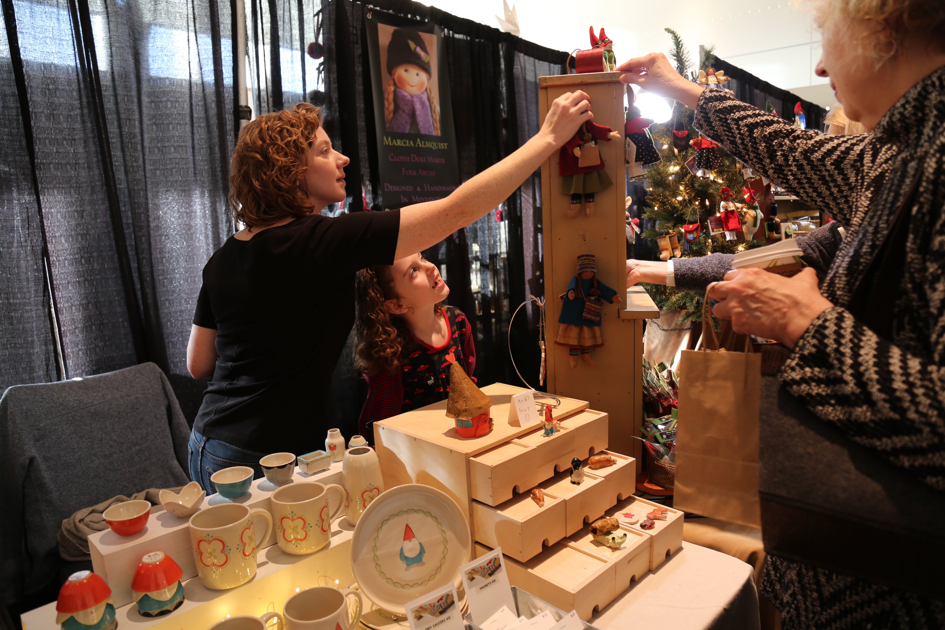 A woman and a child browse handcrafted items at a market stall. The table displays mugs and small decorative items. Another person on the right is reaching towards a wooden display shelf with dolls. A curtain background and festive decor are visible.