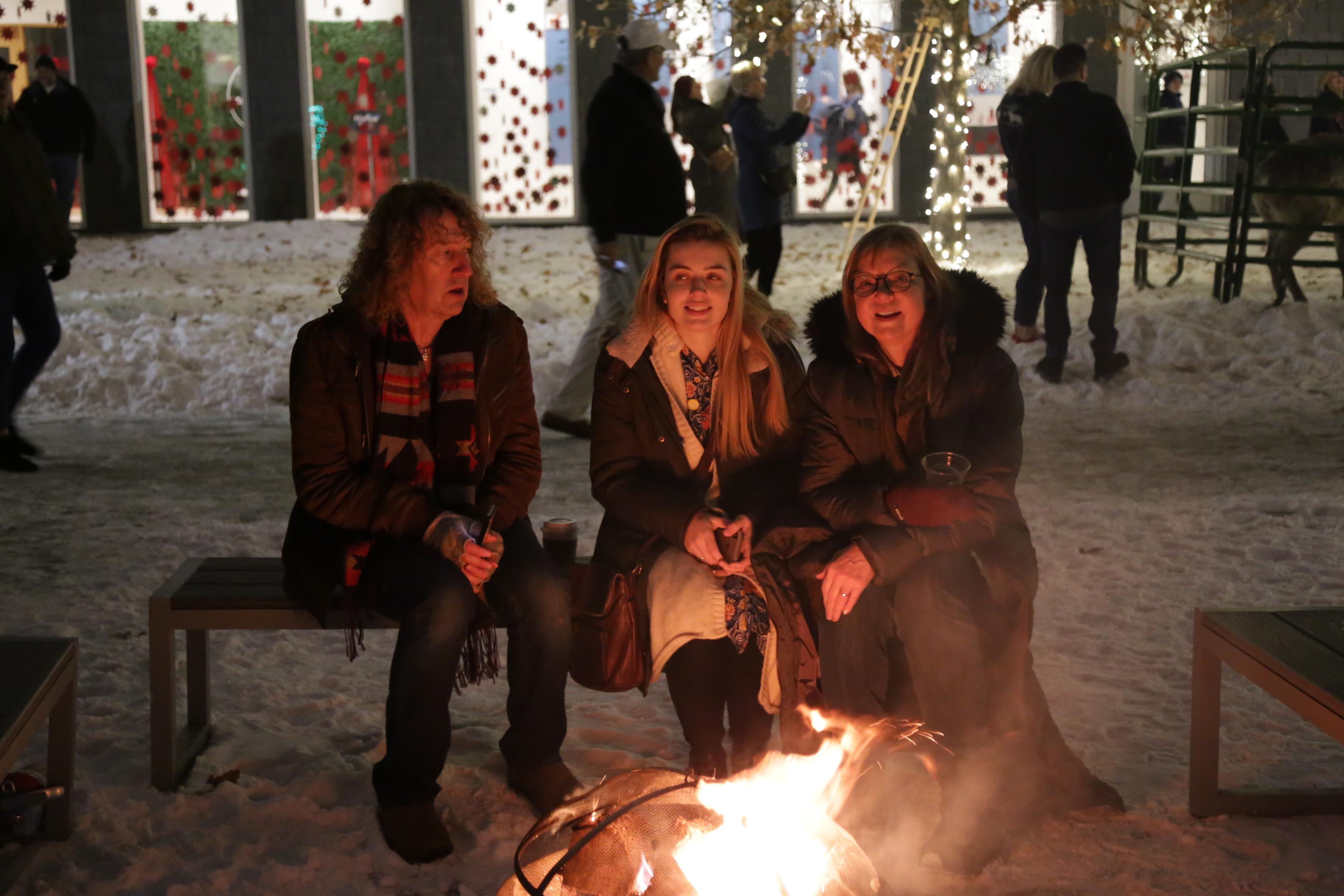 Three people sit around a fire pit outdoors on snowy ground, smiling. Festive lights and decorations are in the background, with people standing and mingling. They are dressed warmly for winter.