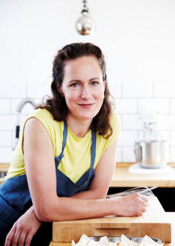 A woman with dark hair, wearing a yellow shirt and blue apron, leans on a wooden kitchen counter with a whisk in her hand. The kitchen has white subway tiles and a stand mixer in the background. She is smiling at the camera.