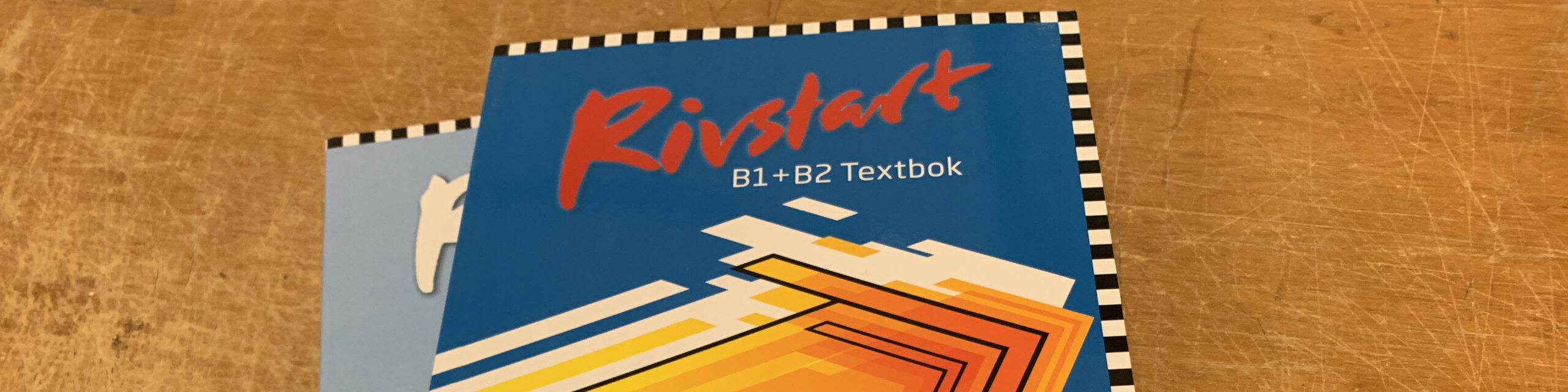 Close-up of the Rivstart B1+B2 Textbok on a wooden surface. The book has a blue cover with geometric orange and yellow designs, and a black-and-white checkered border. A small cat icon is visible on the left.