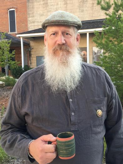 A man with a long white beard wearing a flat cap and a gray shirt holds a striped mug. He stands outdoors in front of a building and trees.